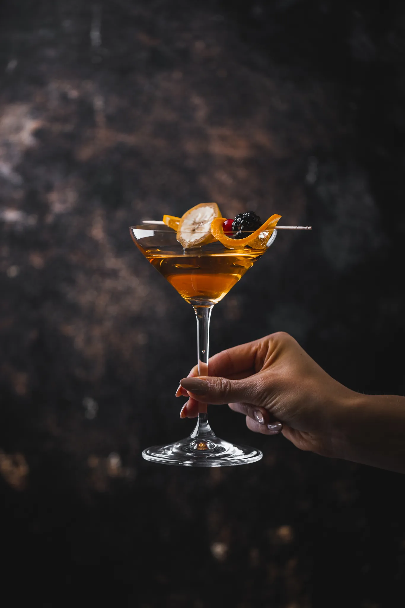 Hand hält einen Cocktail mit Orangenzeste vor dunklem Hintergrund, Restaurant 23 Reasons
