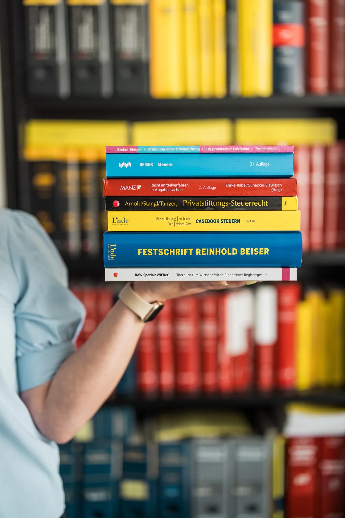 Hand hält einen Stapel Fachliteratur zum Steuerrecht vor der Bibliothek